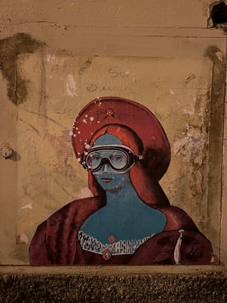 A large-scale wheatpaste artwork on a weathered urban wall depicting a classical Renaissance-style portrait with a surreal twist. The figure features blue skin and is wearing modern scuba goggles, set against a pale, textured background.. Subject: A Renaissance noblewoman with blue skin and a large red headdress, wearing diving goggles.. Style: Painterly and illustrative, blending fine art portraiture with street art subversion.. Details: Intricate lace detailing on the dress, white air bubbles near the goggles, and a signature 'Blub' in the lower right.. Shape: Die-cut portrait bust. Setting: Urban outdoor environment at night.. Surface: A flat utility panel or recessed wall surface with a small keyhole on the left.. Colors: #3388AA (Ocean Blue) , #8B1A1A (Deep Crimson) , #E8D5B5 (Antique Parchment) , #1A1A1A (Carbon Black). Composition: Straight-on, eye-level shot that centers the artwork within the frame.. This piece is part of the 'L'arte sa nuotare' series by artist Blub, which reinterprets iconic artworks as being underwater. It symbolizes the resilience of culture and art, suggesting that even in the face of modern crises or 'floods' of information, classic beauty adapts and survives.