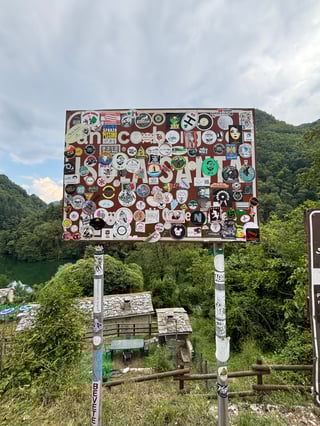 A large brown roadside sign is almost entirely obscured by a dense collage of colorful stickers, set against a backdrop of lush green mountains and a serene lake. The sign serves as a vertical canvas for hundreds of travelers and local groups to mark their presence in this scenic landscape.. Subject: A diverse collection of club logos, travel souvenirs, and brand graphics. Style: Graphic and illustrative collage. Details: The stickers represent various subcultures, from motorcycle clubs like 'Moto Club Alice Castello' to local events and humorous slogans.. Shape: Mixed (circular, rectangular, and die-cut stickers). Setting: Mountainous rural landscape. Surface: An informational or welcome sign on a metal frame with utility poles. Colors: #8B4513 (Saddle Brown), #FFFFFF (White), #2E8B57 (Sea Green), #D3D3D3 (Light Gray). Composition: Straight-on centered composition with a deep depth of field showing the landscape behind the focal point.. This sticker-bombed sign functions as a communal guestbook, where the act of 'slapping' a sticker becomes a ritual of passage for visitors. It highlights a fascinating juxtaposition between the chaotic, ephemeral nature of urban street art and the enduring, static beauty of the natural world.