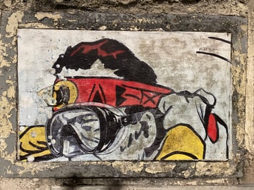 A rectangular wheatpaste artwork is mounted within a recessed frame on a weathered urban wall. It depicts a stylized figure equipped with diving goggles and a red headband, creating a sense of exploration within the city's stone environment.. Subject: A stylized explorer or diver wearing a snorkel mask and a red headband with abstract symbols.. Style: Illustrative and graphic, featuring heavy black outlines, flat color application, and a slightly gritty, hand-finished texture.. Details: The figure sports a red headband with black geometric symbols, yellow elements on the lower sides, and a detailed mask reflecting an abstract grey landscape. A hashtag #ARTESANOGARE is visible in the upper right corner.. Shape: rectangular. Setting: Urban exterior environment.. Surface: A recessed rectangular area on an exterior stone or concrete wall, functioning as a natural frame for the art.. Colors: #ED1C24 (Bright Red), #FFD700 (Golden Yellow), #1A1A1B (Charcoal Black), #D1D3D4 (Light Grey), #808285 (Stone Grey). Composition: Centered and frontal, using the architectural recess of the wall to frame the artwork symmetrically.. The piece uses the metaphor of deep-sea diving to represent urban exploration. By placing a diver on a solid, dry wall, the artist evokes a sense of navigating through the dense 'ocean' of the concrete jungle, seeking hidden truths or simply surviving the pressures of city life.