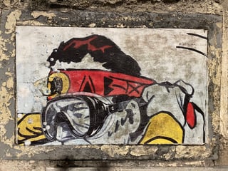 A rectangular wheatpaste artwork is mounted within a recessed frame on a weathered urban wall. It depicts a stylized figure equipped with diving goggles and a red headband, creating a sense of exploration within the city's stone environment.. Subject: A stylized explorer or diver wearing a snorkel mask and a red headband with abstract symbols.. Style: Illustrative and graphic, featuring heavy black outlines, flat color application, and a slightly gritty, hand-finished texture.. Details: The figure sports a red headband with black geometric symbols, yellow elements on the lower sides, and a detailed mask reflecting an abstract grey landscape. A hashtag #ARTESANOGARE is visible in the upper right corner.. Shape: rectangular. Setting: Urban exterior environment.. Surface: A recessed rectangular area on an exterior stone or concrete wall, functioning as a natural frame for the art.. Colors: #ED1C24 (Bright Red), #FFD700 (Golden Yellow), #1A1A1B (Charcoal Black), #D1D3D4 (Light Grey), #808285 (Stone Grey). Composition: Centered and frontal, using the architectural recess of the wall to frame the artwork symmetrically.. The piece uses the metaphor of deep-sea diving to represent urban exploration. By placing a diver on a solid, dry wall, the artist evokes a sense of navigating through the dense 'ocean' of the concrete jungle, seeking hidden truths or simply surviving the pressures of city life.