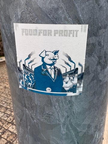 A blue-and-white graphic sticker is centered on a textured metal pole, featuring a socio-political critique of industrial farming. The illustration depicts a pig in a business suit surrounded by livestock and factory smoke.. Subject: Anthropomorphic pig in a suit. Style: Graphic etching-style illustration. Details: The text 'FOOD FOR PROFIT' appears in a blocky font above a pig in corporate attire, with cows, chickens, and smoking factory chimneys in the background.. Shape: Rectangular. Setting: Urban street. Surface: Utility pole. Colors: #004B73 (Deep Blue), #EBEBEB (Off-White), #737D83 (Slate Gray). Composition: Centered, eye-level close-up. The artwork serves as a sharp critique of corporate exploitation in the food industry. By using an anthropomorphic pig in a suit—a classic symbol of greed—it references Orwellian themes to highlight the dehumanization and industrialization of living beings for profit.