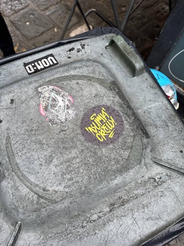 A collection of street art stickers placed on the lid of a weathered, grey plastic trash bin. The most prominent sticker is a circular piece featuring vibrant yellow graffiti-style lettering that reads "'SUMA' CREW!" with a small halo above the 'S'. This sticker has a dark, mottled purple background with a subtle repeating pattern. Adjacent to it is a heavily worn, diamond-shaped sticker with a faded light blue outline and hints of pink, showing significant peeling and distress. A third, rectangular black sticker at the top edge displays the text "VON:D" in a bold, white sans-serif font. The plastic surface of the bin is heavily scratched and dirty, reflecting an gritty outdoor urban environment. In the background, wet cobblestone pavement and metal furniture legs suggest a city street or alleyway setting. The overall mood is one of urban tagging and street-level collective identity.