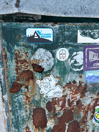 This image captures a collection of diverse stickers, many related to hiking and outdoor activities, adhered to a heavily weathered and rusted green metal utility surface. The contrast between the clean graphics of the stickers and the decaying texture of the metal creates a sense of urban layering and passage of time.. Subject: Mountain climbing and hiking themes. Style: Graphic illustration. Details: Prominent stickers include a rectangular 'Versante Apuano' design with silhouettes of climbers and a die-cut cartoon 'Gamba' hiker character, alongside several smaller round and rectangular tags.. Shape: mixed. Setting: Urban outdoor environment. Surface: Metal utility box or urban infrastructure. Colors: #1E3D31 (Dark Forest Green), #8B4513 (Saddle Brown Rust), #FFFFFF (Pure White), #00AEEF (Cyan Blue). Composition: Close-up, slightly high-angle shot focusing on the cluster of stickers and the surface texture.. The stickers suggest a community of outdoor enthusiasts leaving their mark in an urban environment. The themes of nature and exploration juxtaposed against industrial decay speak to the human desire to bring memories of the wilderness into the urban landscape, creating a physical record of shared subcultural identity.