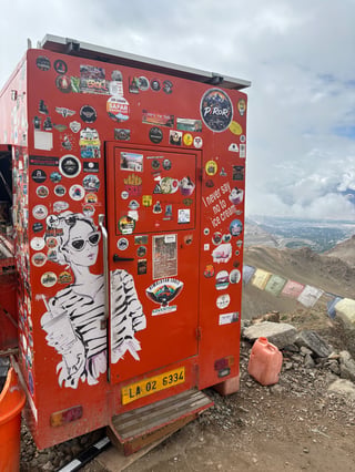A bright red utility unit or mobile shop stands at a high mountain pass, its rear surface transformed into a dense, colorful tapestry of stickers. The centerpiece is a large, stylized illustration of a woman, surrounded by hundreds of smaller travel and lifestyle decals from various adventurers.. Subject: A stylized young woman wearing sunglasses and a striped top, holding a take-away drink cup.. Style: Pop-art sketch style with high-contrast black and white linework.. Details: The figure is flanked by a playful quote 'I never say no to ice cream' and several food-related graphics including fries and a burger.. Shape: Large die-cut. Setting: High-altitude mountain pass in a rugged, arid environment.. Surface: The rear door and body of a mobile utility box or food truck, likely at a high-altitude tourist stop.. Colors: #D32F2F (Crimson Red), #FFFFFF (Pure White), #000000 (Black), #FBC02D (Vibrant Yellow), #5D4037 (Earth Brown). Composition: Vertical framing with a slight low-angle, emphasizing the height of the object and its presence as a landmark against the vast mountain backdrop.. This sticker-covered surface acts as a communal guestbook for the global overlanding and biking subcultures. It represents a collision of modern urban aesthetics—represented by pop-art and commercial decals—and the ancient, spiritual landscape of the Himalayas. Each sticker marks a traveler's presence, turning a functional metal box into a democratic monument of modern adventure.