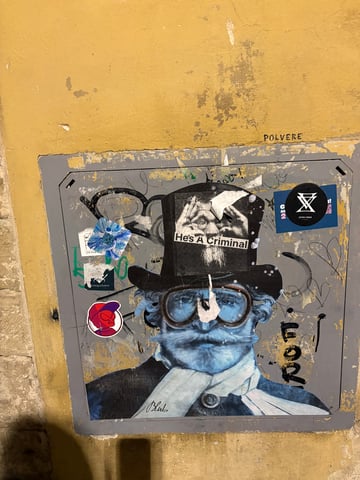 A large wheatpaste artwork by the artist Blub dominates a weathered grey metal hatch set into a textured yellow wall. The piece features a blue-toned gentleman in steampunk attire, overlaid with smaller stickers and urban decay.. Subject: A blue-tinted man with a prominent mustache, goggles, and a top hat.. Style: painterly wheatpaste. Details: The figure is signed 'Blub' at the bottom and has a smaller black and white sticker reading 'He's A Criminal' placed directly on his top hat.. Shape: rectangular. Setting: urban street. Surface: A metal utility access hatch embedded in a plastered wall.. Colors: #3465A4 (Steel Blue), #E9C46A (Ochre Yellow), #2E3436 (Charcoal), #FFFFFF (White). Composition: Centered, frontal shot that emphasizes the rectangular frame of the utility hatch.. This work is part of Blub's 'L'arte sa nuotare' series, which typically portrays figures underwater as a metaphor for resilience during crises. The addition of the 'He's A Criminal' sticker creates a meta-narrative, where the street art itself becomes a surface for further political or social commentary, showcasing the evolving nature of urban dialogue.