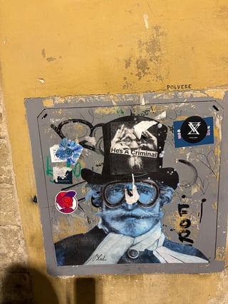 A large wheatpaste artwork by the artist Blub dominates a weathered grey metal hatch set into a textured yellow wall. The piece features a blue-toned gentleman in steampunk attire, overlaid with smaller stickers and urban decay.. Subject: A blue-tinted man with a prominent mustache, goggles, and a top hat.. Style: painterly wheatpaste. Details: The figure is signed 'Blub' at the bottom and has a smaller black and white sticker reading 'He's A Criminal' placed directly on his top hat.. Shape: rectangular. Setting: urban street. Surface: A metal utility access hatch embedded in a plastered wall.. Colors: #3465A4 (Steel Blue), #E9C46A (Ochre Yellow), #2E3436 (Charcoal), #FFFFFF (White). Composition: Centered, frontal shot that emphasizes the rectangular frame of the utility hatch.. This work is part of Blub's 'L'arte sa nuotare' series, which typically portrays figures underwater as a metaphor for resilience during crises. The addition of the 'He's A Criminal' sticker creates a meta-narrative, where the street art itself becomes a surface for further political or social commentary, showcasing the evolving nature of urban dialogue.