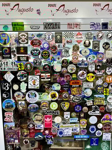 A dense and vibrant collection of stickers covers a transparent glass surface, likely a shop window or display case in a traveler-frequented locale. The stickers are a chaotic mix of graphic styles including bold illustrated logos, minimalist typography, and photographic prints. The predominant themes center around motorcycle culture (numerous 'Moto Clube' emblems), regional tourism across South America (mentions of Ecuador, Atacama, and various Brazilian cities), and sports fandom, particularly Brazilian soccer teams like Cruzeiro E.C. Notable visual elements include a cartoonish chicken drumstick for 'Coxinhas Selvagens', a skull and crossbones for 'Foragidos Moto Clube', and numerous travel-related badges. Colors are eclectic, ranging from bright primary yellows and reds to high-contrast black and white designs. The top of the frame features the repeated red cursive text 'Donde Augusto'. The overall mood is one of a community-driven travel log, capturing the subcultures of South American road trippers, bikers, and urban explorers.