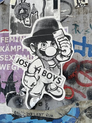 A vertical shot of a layered sticker collage on a gritty urban surface, featuring a central black and white caricature of a video game icon reimagined as a modern hipster.. Subject: A bearded, fedora-wearing version of Mario taking a selfie with a smartphone, holding a 'JOSEPH BOYS' banner.. Style: Illustrated, high-contrast black and white halftone/dot-work.. Details: The character features classic overalls but with a modern, rugged beard and a stylish hat, blending retro gaming culture with contemporary street style.. Shape: die-cut. Setting: Urban street environment. Surface: Urban utility cabinet or metal wall.. Colors: #000000 (Black), #FFFFFF (White), #808080 (Grey), #9B59B6 (Amethyst Purple), #E74C3C (Alizarin Red). Composition: Centered, close-up shot focusing on the main sticker while capturing the surrounding urban context.. The main artwork serves as a playful subversion of pop culture iconography, reimagining a childhood hero as a modern urbanite preoccupied with self-documentation. The 'Joseph Boys' text likely references the artist Joseph Beuys, suggesting a bridge between high art and street subculture. Surrounded by social and political stickers, it represents the dense, multi-vocal nature of urban communication.