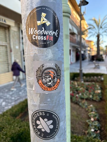 This image captures a weathered, cylindrical metal street pole in an outdoor urban setting, serving as a canvas for local subculture stickers. Three primary stickers are visible: at the top, a circular black sticker for 'Woodwork CrossFit' from Pisa with white and red text; in the middle, a rectangular silver sticker featuring a detailed illustration of a skull wearing an orange motorcycle helmet and goggles, labeled 'FURIOUS TEAM.COM' and 'ITALIE 2024'; and at the bottom, a black and white circular sticker for 'BOARDRIDERS Marina di Pietrasanta'. The setting is a bright, sunlit street scene with a blurred pedestrian, flower beds, and buildings in the background, likely in the Versilia region of Italy. The collection of stickers represents various local communities including fitness, biker culture, and board sports, creating a mosaic of urban identity.