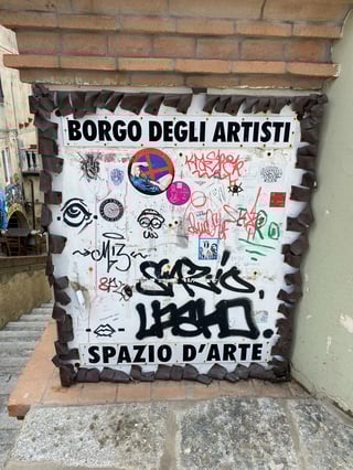 A weathered white metal board, framed by curled rusted metal, serves as a community canvas in a historic Italian setting. Titled 'BORGO DEGLI ARTISTI' and 'SPAZIO D'ARTE', the surface is a dense collage of street culture. It features bold, black spray-painted graffiti tags like 'SERIO' and 'LASH!' alongside delicate hand-drawn icons, including a stylized eye and a face wearing a hat. Numerous stickers are pasted throughout, notably a photographic circular sticker of a woman, a red 'no entry' symbol with a reclining figure, and a political message in German stating 'kein mensch ist illegal' (no human is illegal). The background is filled with various colored markers, scribbles, and the playful text 'PINOCCHIO WAS HERE'. Mounted on a brick and plaster wall next to stone steps, this public art space reflects a vibrant, evolving urban dialogue through its mix of illustration, photography, and typography.