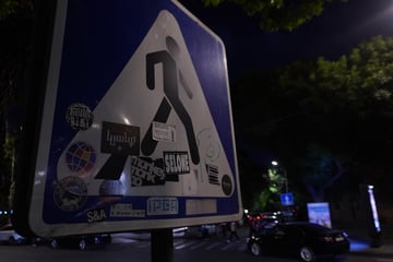 A compilation of diverse urban stickers layered over a blue and white pedestrian crossing sign at night. The composition captures the intersection of official municipal signage and grassroots street culture in an urban setting.. Subject: Graphic logos and typography. Style: Various (typographic, illustrative, and emblem-based). Details: Includes a 'SELONE' tag, an 'FC Pyunik Yerevan' supporter sticker, and Armenian script meaning 'Life'.. Shape: Mixed collection. Setting: Urban night. Surface: Standard pedestrian crossing traffic sign. Colors: #1A3E85 (Deep Blue), #FFFFFF (True White), #000000 (Charcoal Black), #A9A9A9 (Dull Silver). Composition: Low-angle medium shot with a shallow depth of field focusing on the sign's texture.. The sign functions as a democratic canvas, where the rigid authority of the traffic symbol is softened by the chaotic layering of stickers. It represents a living record of the city's subcultures, from local sports fandom to independent artistic tags, showcasing the 'palimpsest' nature of urban surfaces.