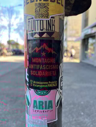 A vertical composition focusing on a metallic pole heavily layered with diverse urban stickers. The central focus is a political sticker promoting solidarity and anti-fascism through the lens of mountain hiking.