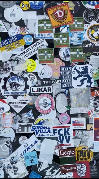 A vibrant and dense 'sticker bomb' collage covering a flat urban surface. The stickers are layered haphazardly, featuring a wide variety of themes including winter sports, football clubs, and travel tags.. Subject: A snowmobile rider silhouette. Style: Graphic logo. Details: The sticker features Russian text 'СНЕГОХОДЧИКИ' (Snowmobilers) in blue and white, set within a stylized gear frame.. Shape: Circular with a gear-like border. Setting: Urban or resort environment. Surface: A flat outdoor utility surface, likely the back of a sign or a control box.. Colors: #FFFFFF (Pure White), #004185 (Deep Blue), #FFD700 (Golden Yellow), #C8102E (Crimson Red), #000000 (Black). Composition: Flat-lay style close-up that fills the entire frame to emphasize the texture and density of the stickers.. This sticker collection serves as a physical guestbook of a specific subculture, likely centered around European alpine tourism and sports. The layering of stickers from different countries (Italy, Germany, Russia) creates a temporal map of visitors, where each layer represents a different moment in time and a different individual leaving their mark on a shared space.