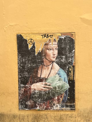 A large-scale wheatpaste artwork applied to a metal utility door, depicting a parody of Leonardo da Vinci's 'Lady with an Ermine'. The central figure is shown holding a large head of lettuce instead of the traditional ermine, set against a vibrant yellow wall.. Subject: A Renaissance-style portrait of a woman holding a head of lettuce.. Style: classical parody. Details: The artwork features significant weathering and peeling at the top. It has been modified with hand-drawn graffiti including an anarchy symbol and the word 'TABOO'.. Shape: rectangular. Setting: urban exterior. Surface: metal utility door embedded in a stucco wall. Colors: #E8C461 (Ochre), #2E2E2E (Charcoal), #7A90A4 (Slate Blue), #889C71 (Sage Green), #7B3625 (Russet). Composition: centered, eye-level frontal shot. This work is a form of 'culture jamming' that subverts high-art iconography to create a surreal and humorous contrast. By replacing the symbol of purity and nobility (the ermine) with a common vegetable, the artist bridges the gap between the elite art world and everyday urban life. The addition of anarchist tagging suggests a rejection of traditional art hierarchies and institutional control.