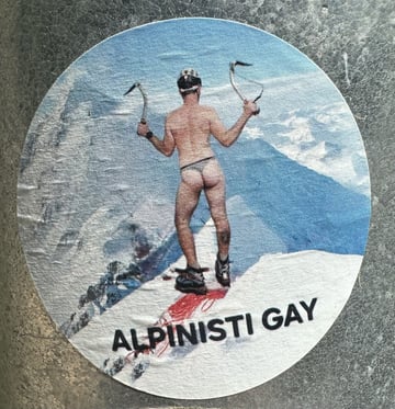 A circular sticker adhered to a metallic surface, featuring a man in a thong climbing a mountain, with the Italian text 'ALPINISTI GAY'. The image combines rugged outdoor imagery with provocative queer visibility.. Subject: A male mountaineer from behind, wearing only a thong, helmet, and boots, holding ice axes.. Style: Photographic with bold typography. Details: The figure is positioned atop a snowy ridge with a red splash of color under the boots. The background shows a crisp blue alpine vista. The text is in a clean, black sans-serif font.. Shape: circular. Setting: Urban outdoor. Surface: A textured metal surface, likely a utility box or a public pole in an urban environment.. Colors: #4A90E2 (Sky Blue), #FFFFFF (Snow White), #000000 (Black), #CC3333 (Deep Red), #B8B8B8 (Steel Grey). Composition: Centered, tight framing that emphasizes the sticker's graphic contrast against the industrial metal background.. This sticker serves as a bold reclamation of space within the mountaineering subculture, which is often perceived as hyper-masculine and heteronormative. By placing a queer-themed image in a public urban space, the artist challenges traditional identities associated with alpinism and promotes LGBTQ+ visibility and pride.
