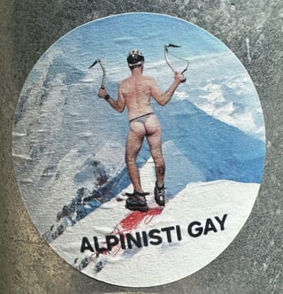 A circular sticker adhered to a metallic surface, featuring a man in a thong climbing a mountain, with the Italian text 'ALPINISTI GAY'. The image combines rugged outdoor imagery with provocative queer visibility.. Subject: A male mountaineer from behind, wearing only a thong, helmet, and boots, holding ice axes.. Style: Photographic with bold typography. Details: The figure is positioned atop a snowy ridge with a red splash of color under the boots. The background shows a crisp blue alpine vista. The text is in a clean, black sans-serif font.. Shape: circular. Setting: Urban outdoor. Surface: A textured metal surface, likely a utility box or a public pole in an urban environment.. Colors: #4A90E2 (Sky Blue), #FFFFFF (Snow White), #000000 (Black), #CC3333 (Deep Red), #B8B8B8 (Steel Grey). Composition: Centered, tight framing that emphasizes the sticker's graphic contrast against the industrial metal background.. This sticker serves as a bold reclamation of space within the mountaineering subculture, which is often perceived as hyper-masculine and heteronormative. By placing a queer-themed image in a public urban space, the artist challenges traditional identities associated with alpinism and promotes LGBTQ+ visibility and pride.