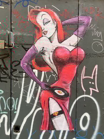 A large, hand-drawn wheatpaste of Jessica Rabbit applied to a weathered grey metal door covered in graffiti. The character is depicted in her signature seductive pose but with a domestic twist, holding a frying pan with an egg.. Subject: A stylized Jessica Rabbit holding a frying pan. Style: Illustrated, hand-painted with markers and bold line work. Details: Iconic red dress and purple gloves, featuring a Superman logo on a black garter belt and a sunny-side-up egg in a pan.. Shape: Large, die-cut silhouette. Setting: Urban street environment. Surface: Metal utility door or service cabinet. Colors: #E6192E (Vibrant Red), #8C4E9C (Deep Purple), #333333 (Charcoal Grey), #FFFFFF (Pure White), #F7B500 (Golden Yellow). Composition: Vertical full-body shot centered on the artwork. This piece by artist #LeDieSis subverts the traditional femme fatale archetype by placing Jessica Rabbit in a domestic context. The juxtaposition of her overt sexuality with a mundane task like frying an egg provides a humorous commentary on gender roles, while the Superman logo adds a layer of pop-culture empowerment.