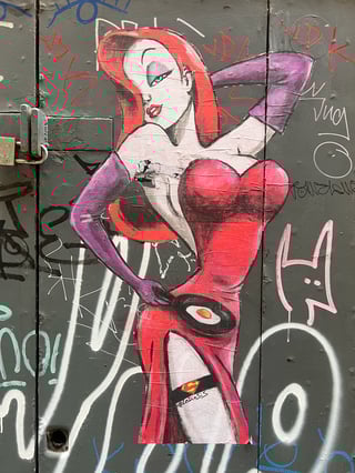 A large, hand-drawn wheatpaste of Jessica Rabbit applied to a weathered grey metal door covered in graffiti. The character is depicted in her signature seductive pose but with a domestic twist, holding a frying pan with an egg.. Subject: A stylized Jessica Rabbit holding a frying pan. Style: Illustrated, hand-painted with markers and bold line work. Details: Iconic red dress and purple gloves, featuring a Superman logo on a black garter belt and a sunny-side-up egg in a pan.. Shape: Large, die-cut silhouette. Setting: Urban street environment. Surface: Metal utility door or service cabinet. Colors: #E6192E (Vibrant Red), #8C4E9C (Deep Purple), #333333 (Charcoal Grey), #FFFFFF (Pure White), #F7B500 (Golden Yellow). Composition: Vertical full-body shot centered on the artwork. This piece by artist #LeDieSis subverts the traditional femme fatale archetype by placing Jessica Rabbit in a domestic context. The juxtaposition of her overt sexuality with a mundane task like frying an egg provides a humorous commentary on gender roles, while the Superman logo adds a layer of pop-culture empowerment.