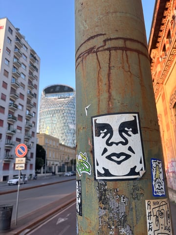 A close-up shot of a weathered green utility pole adorned with several street stickers, most notably the iconic black and white Andre the Giant face. The pole's rusted texture contrasts with the modern glass-and-steel architecture of a skyscraper visible in the blurred background.. Subject: The stylized face of Andre the Giant, famously known as the 'OBEY' icon.. Style: High-contrast, stencil-like graphic illustration.. Details: The sticker shows some wear, with the bottom-right corner slightly peeling away from the rusted surface.. Shape: rectangular. Setting: Urban cityscape in Milan, featuring modern and traditional architecture.. Surface: A cylindrical metal utility or lamp pole positioned on an urban sidewalk.. Colors: #000000 (Black), #FFFFFF (White), #8B4513 (Saddle Brown), #8A9A5B (Sage Green), #87CEEB (Sky Blue). Composition: Macro-style focus on the sticker with a shallow depth of field, using the pole as a vertical lead-in to the blurred urban backdrop.. The presence of the 'OBEY' sticker—a symbol of Shepard Fairey's phenomenology movement—serves as a reminder to question the 'visual landscape' of the city. Placed on a decaying piece of infrastructure in front of a gleaming corporate tower, it highlights the juxtaposition between grassroots street culture and the polished facade of modern urban development.