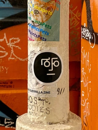 A close-up shot of a weathered utility pole in an urban environment, heavily adorned with stickers and graffiti. The focal point is a circular black sticker with a white minimalist graphic that resembles a stylized, geometric face. Above it, a sticker for 'PaintsPets' displays a cat illustration alongside the brand name repeated in green, blue, and yellow. Below the central black sticker is another label referencing 'URBAN GUERRILLA ZINE' and '9/11', while 'LOST BODIES MUSIC' is hand-written directly onto the pole in dark ink. The setting is vibrant and gritty, with a background of bright orange walls covered in white graffiti tags and scribbles. This composition captures the layers of DIY culture, from zines and music to animal-themed art, within a bustling city landscape.