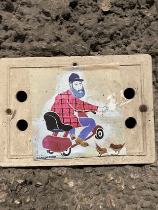 A rectangular sticker featuring a whimsical illustration of a bearded man riding a small motorized tricycle. The artwork uses a flat, illustrative style and is placed on a utility panel against a rough, textured wall.