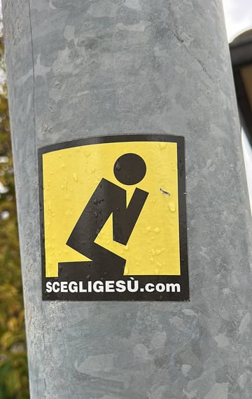 A square religious sticker featuring a minimalist black pictogram of a person kneeling in prayer, set against a vibrant yellow background. The design is reminiscent of a cautionary or informational sign, framed by a thick black border. At the bottom, white text on a black bar reads 'SCEGLIGESU.COM', which translates from Italian as 'Choose Jesus'. The sticker is applied to a grey, galvanized metal pole, likely a utility post or lamppost, and is covered in small water droplets from recent rain. The blurry background suggests an outdoor urban or suburban setting with hints of green and yellow foliage. This piece serves as a form of religious outreach or street mission, using the visual language of urban signage to convey a spiritual message.