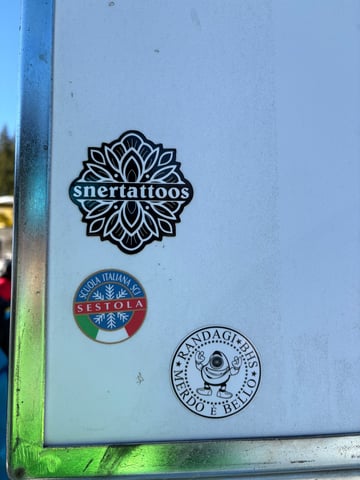 A collection of three distinct stickers on a white metal surface, likely part of a utility box or vehicle in an outdoor setting. The top sticker features a high-gloss, black and white mandala-inspired floral pattern with the text 'snertattoos' in a central band. Below it is a colorful circular emblem for 'SCUOLA ITALIANA SCI SESTOLA', showcasing blue, red, and the Italian flag colors with stylized white snowflakes. The bottom-most sticker is a black and white circular design featuring an illustrated one-eyed cyclops monster character making rock-and-roll hand gestures, surrounded by the Italian text 'RANDAGI BHS' and 'MERDO È BELLO' with small stars. This eclectic mix captures a blend of professional sports branding, tattoo artistry, and underground street culture, set against a clean, industrial background under bright daylight.