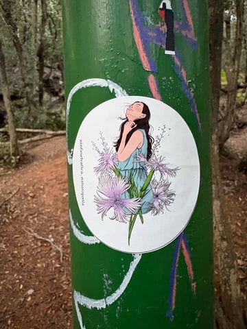 An oval-shaped illustrated sticker featuring a woman with dark hair and her head tilted back in a gesture of peaceful ecstasy. She wears a light blue sleeveless dress and is surrounded by large, delicately fringed flowers in shades of pale purple and white, resembling wild dianthus. The style is a clean, modern illustration with soft, watercolor-like shading. The text 'ninfaselvatica_naturopatia' is printed vertically along the left edge. The sticker is placed on a weathered green metal pole, set against the backdrop of a forest trail with trees and a dirt path. The artwork conveys a theme of natural healing, serenity, and a deep connection to the wilderness.