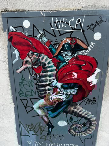 A large-scale wheatpaste parodying Jacques-Louis David's 'Napoleon Crossing the Alps', depicting a military figure riding a giant seahorse. The artwork is applied to a heavily tagged metal utility box, creating a contrast between the detailed illustration and raw urban graffiti.. Subject: A figure resembling Napoleon Bonaparte in a bicorne hat and flowing red cape, riding a large, realistic seahorse.. Style: Painterly illustration with bold outlines. Details: The figure wears modern-looking goggles and the seahorse features intricate brown and gray striped patterns.. Shape: Die-cut. Setting: Urban exterior. Surface: A metal utility box or electrical cabinet. Colors: #C41E3A (Cardinal Red), #2B4F60 (Deep Teal), #D6C5B3 (Parchment), #000000 (Black), #FFFFFF (White). Composition: Straight-on centered shot highlighting the scale of the artwork relative to the utility box.. This piece subverts classical iconography by replacing the traditional warhorse with a seahorse, blending historical grandeur with surrealist humor. It serves as a commentary on the fluidity of power and the recontextualization of 'high art' within the public, urban sphere.