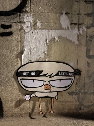 A large, stylized die-cut sticker of a grumpy bird character holding a baseball is affixed to a weathered urban wall. The character wears a black headband with the iconic Ramones lyrics 'HEY HO LET'S GO'.. Subject: A stylized bird character with large white eyes, an orange beak, and a punk-rock headband holding a baseball.. Style: Comic-inspired pop art using halftone Ben-Day dots for shading and texture.. Details: The headband features the text 'HEY HO LET'S GO' in a bold, sans-serif font, and the bird has a determined or grumpy expression.. Shape: Die-cut silhouette following the character's form. Setting: Urban street environment. Surface: An urban wall, possibly in an alleyway or subway passage, showing signs of layering and age.. Colors: #FFFFFF (White), #000000 (Black), #E89E62 (Muted Orange), #C4BFA6 (Stone Beige). Composition: A centered, medium close-up shot that emphasizes the sticker's contrast against the rough texture of the wall.. The piece bridges the gap between sports culture and punk rock subculture. By referencing The Ramones' 'Blitzkrieg Bop' and using a comic-book halftone aesthetic, the artist creates a sense of urban grit mixed with playful rebellion. The character's defiant stance reflects the typical 'don't care' attitude of the street art scene.