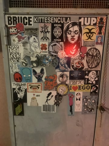 A dense sticker bomb covers the upper portion of a grey metal door, showcasing a wide variety of styles and artists. The collection ranges from bold typography and street tags to intricate character illustrations and graphic icons.. Subject: A stylized feminine face with dark hair and hollow eyes. Style: Graphic noir illustration. Details: The sticker features heavy black ink lines and is caught in a strong red glare from a nearby light source, emphasizing the lips and chin.. Shape: die-cut rectangular. Setting: Urban environment. Surface: A metal utility door or electrical cabinet, likely located in an urban alley or transit hub.. Colors: #000000 (Jet Black), #FFFFFF (Cloud White), #FF0000 (Glowing Red), #808080 (Industrial Grey), #E9C46A (Ochre Yellow). Composition: Straight-on, medium close-up shot that captures the density and layering of the stickers.. This sticker bomb functions as a collaborative palimpsest, where individual artists like 1UP and Bruce reclaim public infrastructure. It represents the chaotic, democratic nature of street art, where disparate voices, political slogans, and personal mascots converge into a singular, evolving visual narrative of the city's subculture.