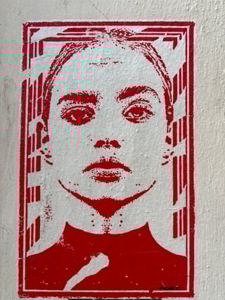 A striking red and white wheatpaste poster featuring a stylized, high-contrast portrait of a woman. The artwork is affixed to a textured, off-white urban wall, showcasing the tactile wrinkles of manual application.. Subject: A portrait of a woman with an intense, steady gaze. Style: High-contrast stencil or screen-print aesthetic. Details: The portrait is enclosed in a decorative frame with geometric patterns and vertical bars. The bottom right features a small signature that appears to read 'ACHES'.. Shape: Rectangular. Setting: Urban environment. Surface: Exterior building wall. Colors: #C41E3A (Cardinal Red), #FFFFFF (White), #E2DED0 (Almond). Composition: A centered, eye-level close-up shot that emphasizes the symmetry and intensity of the portrait.. The bold red ink and high-contrast style create a sense of urgency and authority, reminiscent of classic propaganda posters but repurposed for urban expression. By placing a large-scale portrait in a public space, the artist humanizes the concrete environment, transforming a silent wall into a platform for presence and identity.