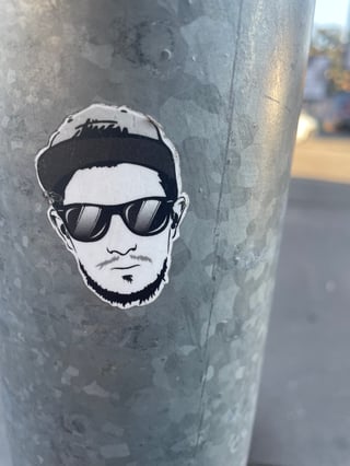 A high-contrast black and white sticker of a man's face is affixed to a weathered galvanized metal pole. The artwork stands out against the textured metallic surface, capturing a moment of urban street life.