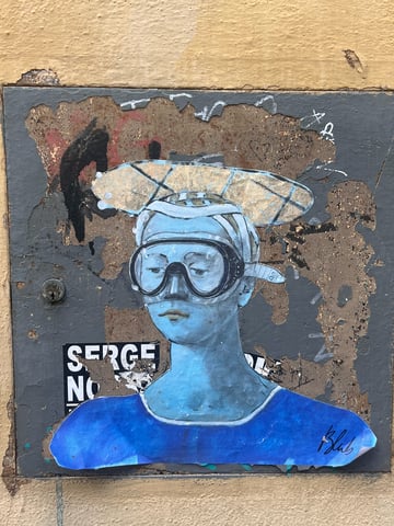 A blue-tinted wheatpaste artwork depicting a classical-style portrait of a figure wearing modern diving goggles. The piece is pasted onto a weathered, metallic utility door that shows significant signs of age and decay.. Subject: A person in Renaissance attire with a scuba mask. Style: Illustrated portrait blending classical and contemporary elements. Details: The figure features monochromatic blue skin tones, a detailed diving mask with glass reflections, and a large patterned headdress. The signature 'Blub' is visible in the lower right.. Shape: die-cut. Setting: Urban street level. Surface: A metal utility panel or access door. Colors: #2E74E5 (Azure Blue), #BDB76B (Dark Khaki), #555555 (Dark Grey), #D2B48C (Tan). Composition: A centered, eye-level portrait shot that captures the interaction between the clean lines of the art and the gritty texture of the surface.. Part of the 'L'arte sa nuotare' (Art knows how to swim) series by Blub, this work recontextualizes iconic classical art within a modern, aquatic metaphor. By placing a diving mask on the subject, the artist suggests that art is resilient and can survive the 'floods' of time and changing social landscapes.