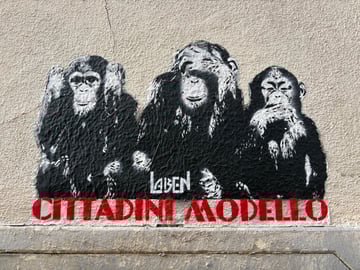 A high-contrast black and white stencil of the three wise monkeys is depicted on a textured urban wall, accompanied by bold red text in Italian. The piece combines traditional iconography with contemporary political irony.. Subject: Three chimpanzees illustrating the 'See No Evil, Hear No Evil, Speak No Evil' maxim.. Style: Multi-layered high-contrast stencil art. Details: The monkeys are rendered with textured shading; beneath them is the text 'CITTADINI MODELLO' in a red industrial stencil font and the artist's tag 'LAIBEN'.. Shape: Rectangular stencil composition. Setting: Urban exterior. Surface: An exterior building wall, positioned just above a concrete foundation ledge.. Colors: #000000 (Black), #FFFFFF (White), #C61A1E (Ruby Red), #E8E1D5 (Sandstone). Composition: Straight-on, centered shot that captures the symmetry of the figures and the horizontal flow of the text.. This work uses the 'Three Wise Monkeys' to satirically redefine the concept of a 'Model Citizen' (Cittadini Modello). It suggests that the ideal citizen from a systemic perspective is one who remains willfully ignorant or silent regarding social and political corruption, turning a traditional moral proverb into a critique of civic apathy.