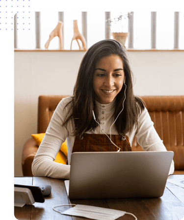 Smiling woman on a video call wearing earphones and working on a laptop in a cozy, well-lit workspace.