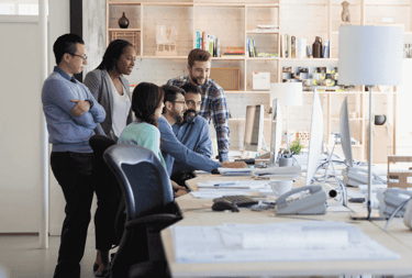 Coworkers gathered around a desk collaborating and reviewing work on a computer in a modern open office space