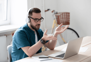 Remote worker wearing headphones gesturing during a video call on laptop at home office desk.