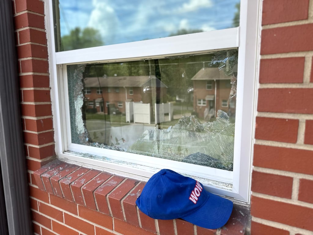 Broken residential window with shattered glass pane in white frame, blue WOW cap on brick windowsill, showing repair need