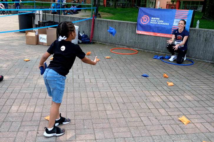 A Rising New York Road Runners student at a bean bag toss station with Jenny Simpson