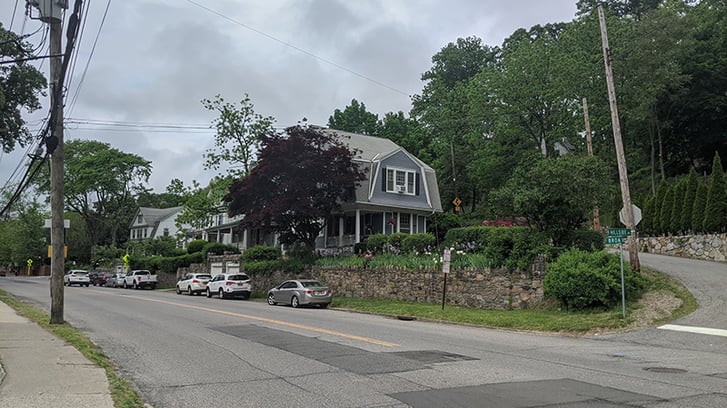 A wide street in the suburbs with a blue house on the corner and lots of trees.