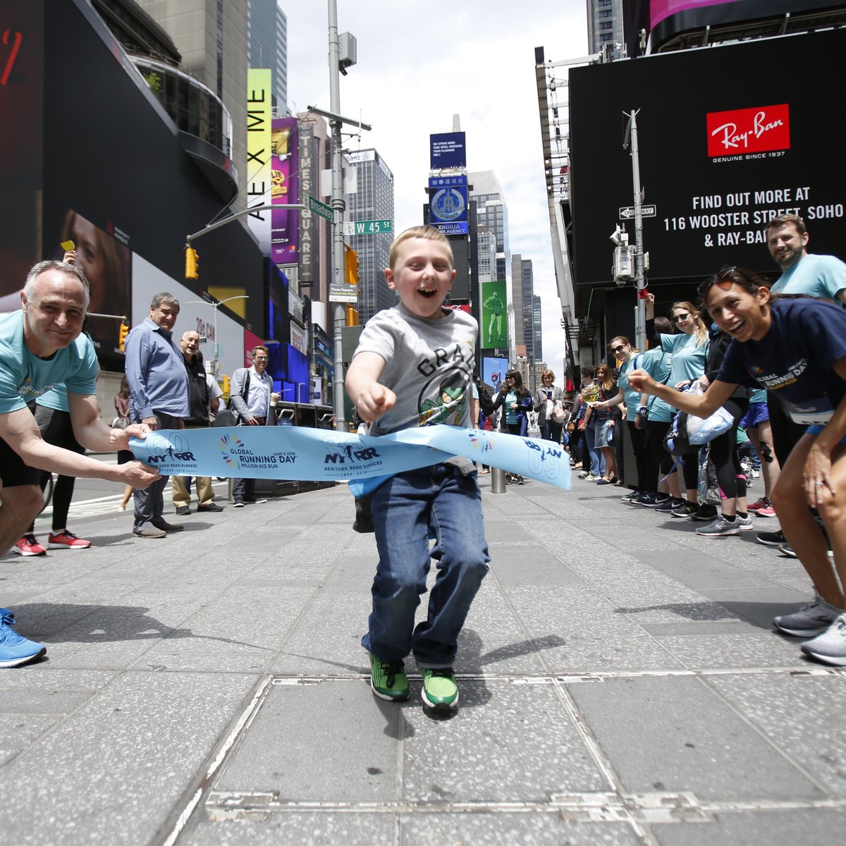 Des Linden and NYRR CEO Michael Capiraso hold a pop-up finish line tape in Times Square as a young runner crosses through to celebrate Global Running Day.