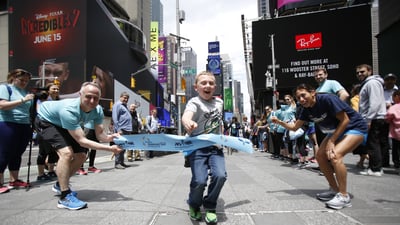 Des Linden and NYRR CEO Michael Capiraso hold a pop-up finish line tape in Times Square as a young runner crosses through to celebrate Global Running Day.