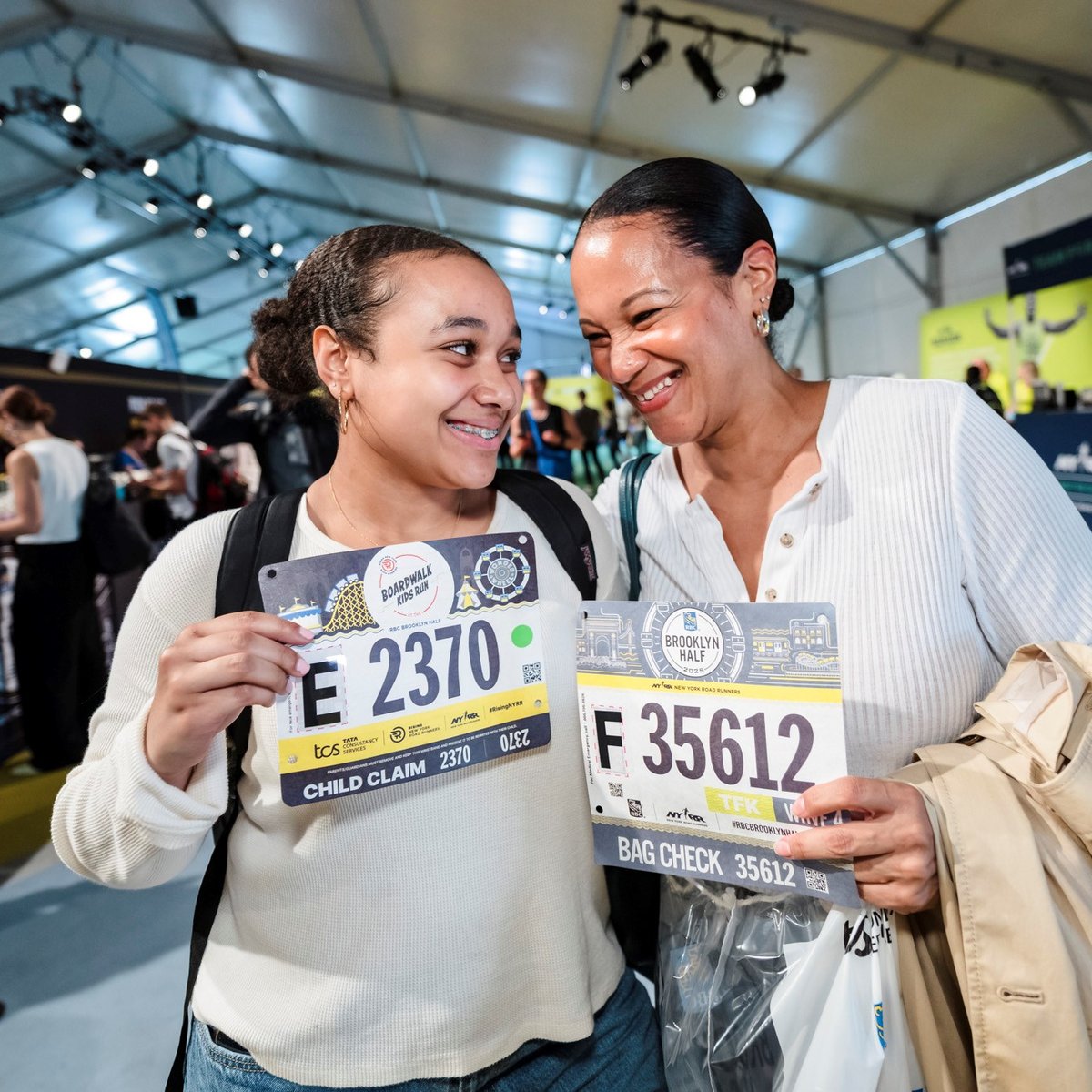 A mom and daughter smile at each other holding their bibs at the 2025 RBC Brooklyn Half Pre-Party