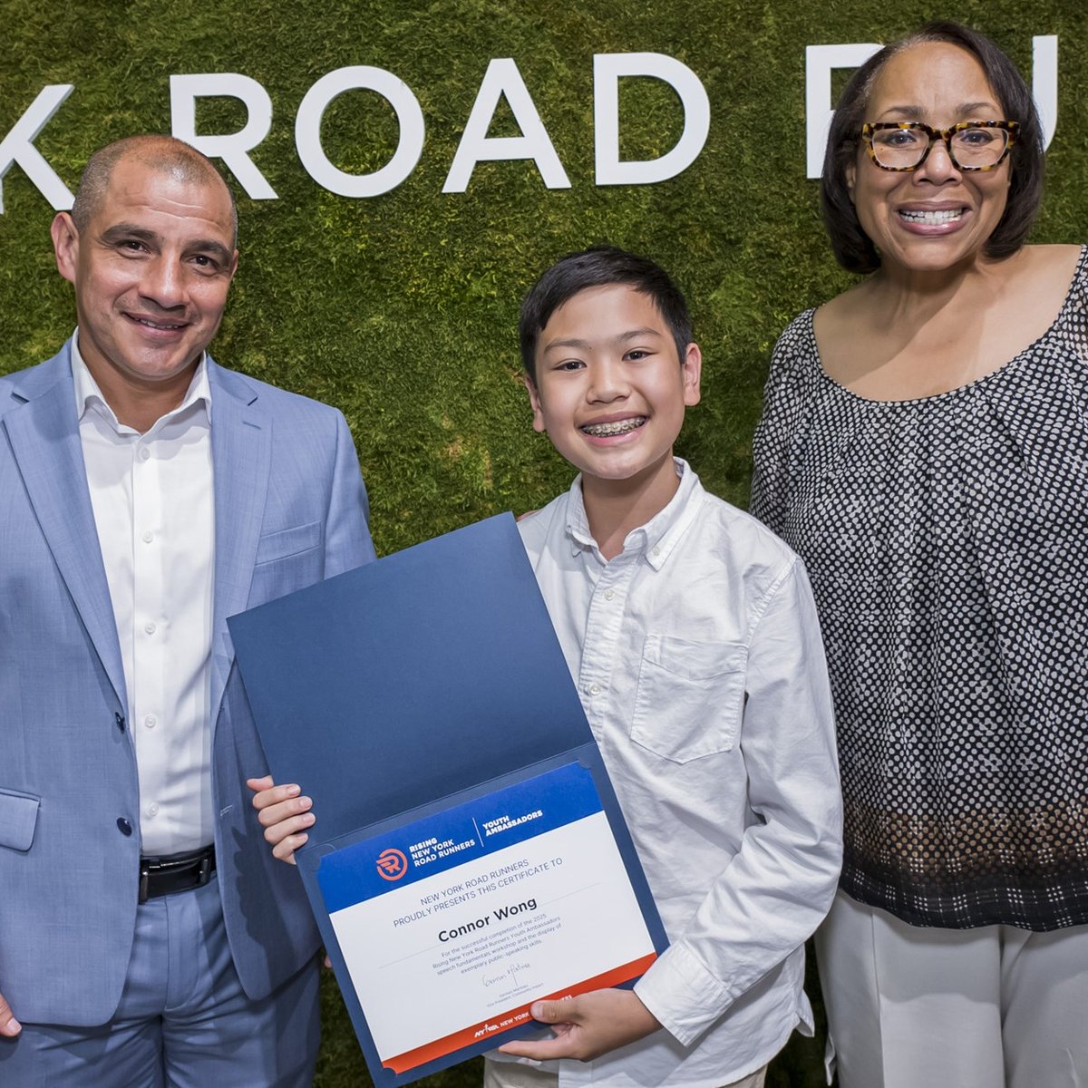 A youth ambassador smiles with his certificate and two NYRR employees at graduation