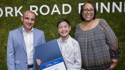 A youth ambassador smiles with his certificate and two NYRR employees at graduation