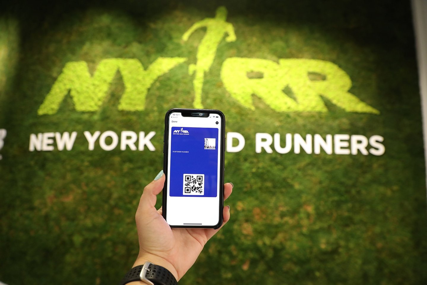 A hand holding a phone in front of the NYRR RUNCENTER's green wall; the phone's screen shows a QR code