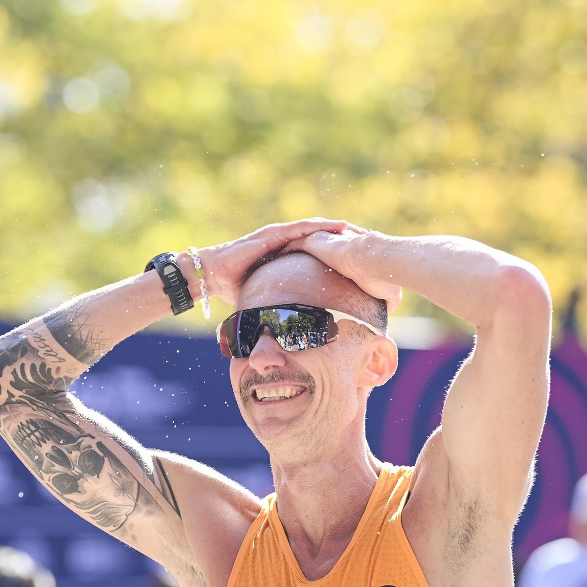 Male runner with a mustache sunglasses and skull tattoo on his bicep puts his hands on his head and smiles after crossing the finish line at the TCS New York City Marathon