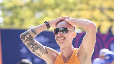Male runner with a mustache sunglasses and skull tattoo on his bicep puts his hands on his head and smiles after crossing the finish line at the TCS New York City Marathon
