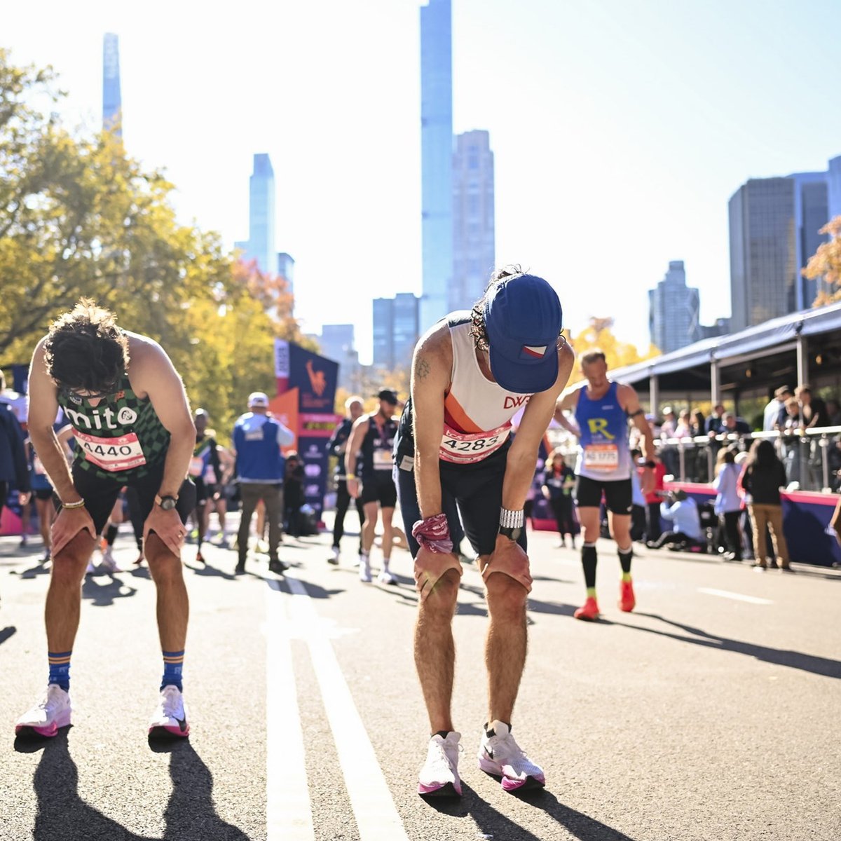 Two runners bent over with their hands on their knees with the TCS New York City Marathon finish line and other runners behind them